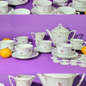 Könnte beinhalten: Ein weißes Porzellan-Teeservice mit Goldrand und floralen Akzenten. Das Set umfasst eine Teekanne, einen Milchgießer, eine Zuckerdose, Tassen und Untertassen. Das Teeservice steht auf einer lila Oberfläche.