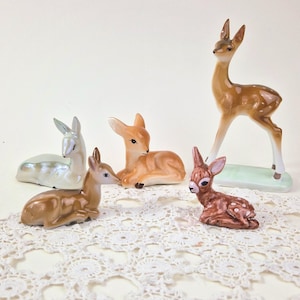 May include: A collection of ceramic deer figurines in various poses. The deer are in shades of brown, tan, and pearlescent white. One deer is standing, while the others are lying down. The figurines are on a white lace surface.