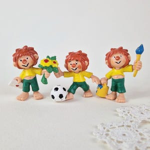 May include: Three small figurines of characters with messy orange hair, yellow shirts, and green shorts. The first holds a bouquet of yellow flowers and a white envelope. The second kicks a black and white soccer ball. The third holds a blue paintbrush and a yellow paint bucket.