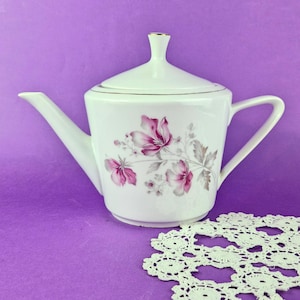 Puede incluir: Una tetera de porcelana blanca con un delicado estampado floral de flores rosas y grises en el lateral. La tetera incluye una tapa a juego con un pequeño pomo, un pico curvo y un asa resistente, todo ello rematado con un fino borde dorado.