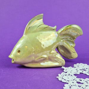 Könnte beinhalten: Eine Keramikfischfigur mit einer glänzenden, irisierenden hellgrünen Oberfläche. Der Fisch hat eine ausgeprägte Rückenflosse, eine breite Schwanzflosse und ein kleines kreisförmiges Auge. Die Oberfläche reflektiert das Licht.