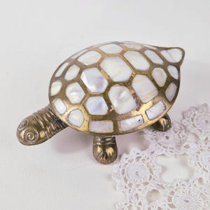 Könnte beinhalten: Eine dekorative Schildkrötenfigur aus Messing mit polierter Oberfläche. Der Panzer ist mit einem Mosaikmuster aus schillernden, weißen Perlmuttsegmenten eingelegt. Kopf, Beine und Schwanz bestehen aus massivem Messing mit Gravuren.