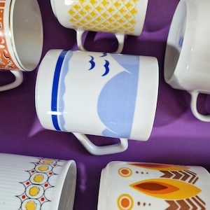 Może przedstawiać: Kolekcja białych ceramicznych kubk&oacute;w z r&oacute;żnymi wzorami. Jeden kubek ma niebiesko-biały wz&oacute;r żagl&oacute;wki z trzema niebieskimi ptakami. Inne kubki mają ż&oacute;łte, pomarańczowe i brązowe geometryczne wzory.