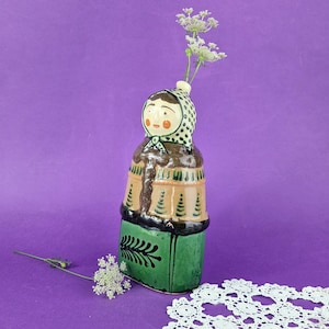 Può includere: Un vaso in ceramica a forma di persona che indossa un foulard a pois, un cappotto marrone con motivi di alberi verdi e una gonna verde con un disegno di foglie nere. Un piccolo rametto di fiori bianchi è inserito nella parte superiore.