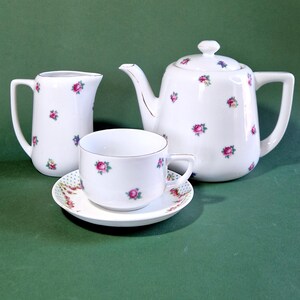 Könnte beinhalten: Ein weißes Porzellan-Teeservice mit Teekanne, Sahnekännchen, Tasse und Untertasse. Jedes Teil ist mit kleinen rosa und lila Blumenmustern verziert. Die Teekanne hat einen gebogenen Ausguss und Henkel.