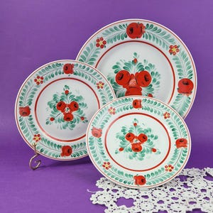 Op de afbeelding: Een set van drie witte keramische borden met een bloemenontwerp. Elk bord heeft een rode rand, een centrale opstelling van rode bloemen en een groene en rode bladrand. De borden zijn geplaatst op een paarse ondergrond.