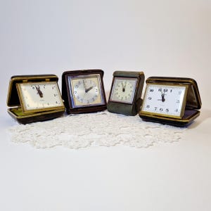 May include: Four vintage alarm clocks with black leather cases and silver faces. The clocks are arranged in a row on a white lace doily.