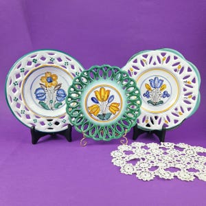 May include: Three decorative ceramic plates with floral designs. The plates feature a white background with blue, yellow, and green accents. The plates are displayed on black stands against a purple background. One plate has a green openwork border.