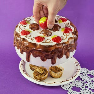Può includere: Un contenitore in ceramica a forma di torta con base bianca, glassa al cioccolato e decorazioni di fragole rosse. Il contenitore è posto su un piatto bianco con tre muffin.