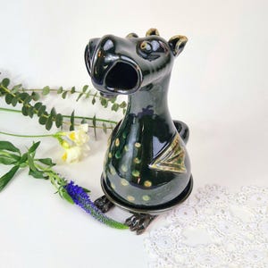 Può includere: Una statuetta di drago in ceramica verde scuro con finitura lucida, caratterizzata da una bocca spalancata, piccole ali sui lati e macchie circolari verde chiaro sul corpo arrotondato. Il drago poggia su una base circolare coordinata.
