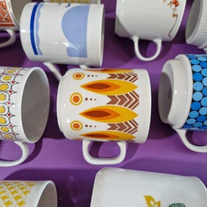Może przedstawiać: Kolekcja białych ceramicznych kubk&oacute;w z r&oacute;żnymi kolorowymi wzorami. Wzory obejmują kształty geometryczne, motywy kwiatowe i sztukę abstrakcyjną w odcieniach ż&oacute;łci, pomarańczy, niebieskiego i brązu. Każdy kubek ma zakrzywiony uchwyt.