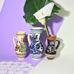 May include: Three ceramic pitchers with handles, each featuring unique hand-painted designs. The pitchers are in cream, blue, black, and orange, with floral and abstract patterns. A white calla lily and green leaf are in the background.