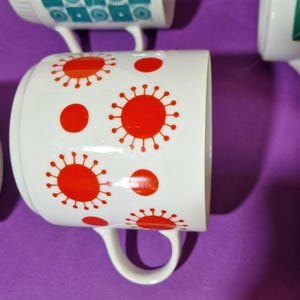 Może przedstawiać: Białe ceramiczne kubki z czerwonymi wzorami promieni słonecznych i kropek. Kubki mają zakrzywiony uchwyt i są ustawione na fioletowym tle. Kubki są częścią zestawu.
