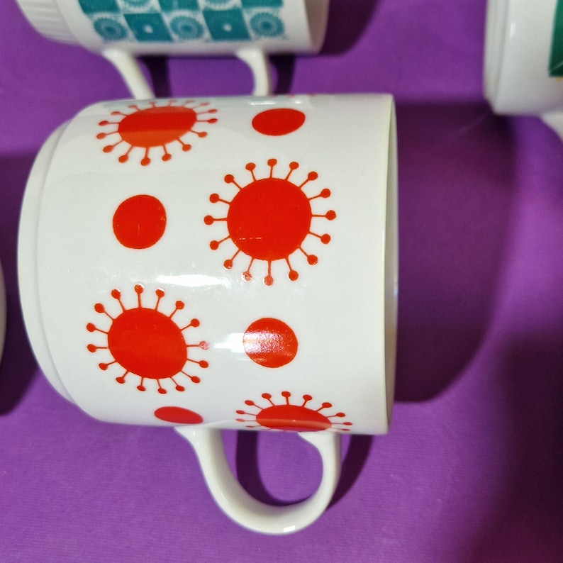 Może przedstawiać: Białe ceramiczne kubki z czerwonymi wzorami promieni słonecznych i kropek. Kubki mają zakrzywiony uchwyt i są ustawione na fioletowym tle. Kubki są częścią zestawu.