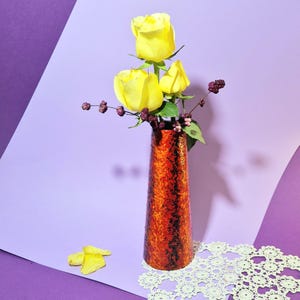 Può includere: Un vaso arancione con superficie strutturata contiene tre rose gialle e rametti di bacche. Il vaso è posto su una tovaglietta di pizzo bianco, su sfondo viola. Petali gialli sparsi vicino al vaso.