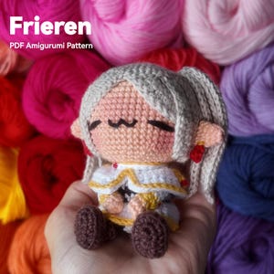 May include: A crocheted amigurumi doll of a character with gray hair and a white and gold outfit. The doll is labeled "Frieren PDF Amigurumi Pattern". The background features colorful yarn skeins in shades of pink, red, and purple.