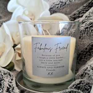 May include: A clear glass candle jar with a white candle inside. The label on the jar reads "Fabulous Friend" and includes a message about friendship. The candle is sitting on a silver base.