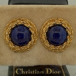 Könnte beinhalten: Ein Paar runde Clip-Ohrringe mit einem tiefblauen, marmorierten Cabochon-Zentrum, umgeben von einem strukturierten, geflochtenen goldfarbenen Metallrand. Die Ohrringe befinden sich in einem schwarzen Etui mit dem goldenen Schriftzug Christian Dior.