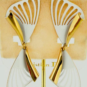Könnte beinhalten: Ein Paar weiß-goldfarbene Ohrringe. Die Ohrringe haben ein flügelförmiges Design mit weißer Emaille-Oberfläche und goldfarbenen Akzenten. Die Ohrringe werden auf einem beigen Hintergrund präsentiert.