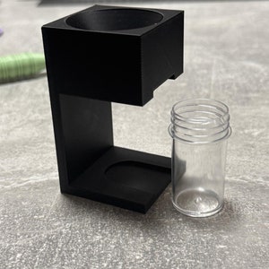 Op de afbeelding: Een zwarte 3D-geprinte houder met een ronde opening aan de bovenkant en een kleinere opening aan de zijkant. Naast de houder bevindt zich een doorzichtige plastic buis.