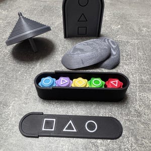 Könnte beinhalten: Ein Satz 3D-gedruckter Spielsteine, bestehend aus einem Kreisel, einem Tor, zwei Steinen, einer Box mit sechs farbigen Knöpfen und einem rechteckigen Stück mit Symbolen. Die Symbole sind ein Quadrat, ein Dreieck und ein Kreis.