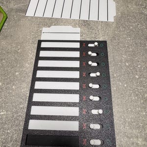 Op de afbeelding: Een zwart-wit scorebord met een reeks witte gleuven en een rij witte schakelaars. Elke gleuf heeft een rode 'X' en een groene cirkel. Het bord heeft een zwart gedeelte aan de onderkant.
