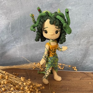 Könnte beinhalten: Gehäkelte Medusa-Puppe mit grünen Schlangenhaaren, einem hellbraunen Gesicht und einem gelb-grünen Kleid. Die Puppe hat schwarze Augen und steht auf einer Holzoberfläche. Das Haar der Puppe hat rote Akzente.