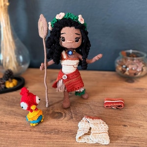 May include: A crocheted doll of Moana from the Disney movie Moana, wearing a brown and orange skirt, a white top, and a flower crown. The doll is holding a wooden stick and is standing on a wooden surface. There is a small crocheted red and yellow bird and two pieces of crocheted clothing next to the doll.