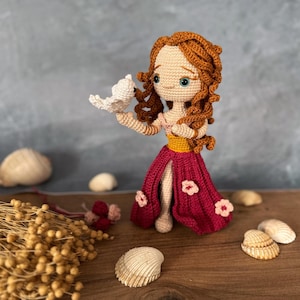 May include: A crocheted doll with long, curly red hair wearing a burgundy dress with pink flowers. The doll is holding a white crocheted bird in its hand.