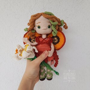 May include: Crocheted doll with orange and yellow wings, brown hair, and a green flower in its hand. The doll is wearing a red dress and green shoes. The doll is holding a white flower.