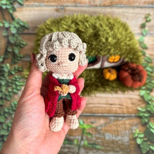 May include: A crocheted doll with blonde hair, wearing a red coat and holding a small brown object. The doll is standing in a hand, with a green background.