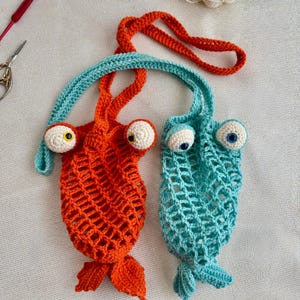 May include: Two whimsical, handmade crochet fish-shaped bags. One is bright orange with a blue handle and the other is turquoise with an orange handle. Both have large, googly eyes with yellow and blue centers, and a fish tail.