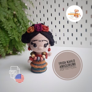 Może przedstawiać: Lalka amigurumi wykonana na szydełku przedstawiająca Fridę Kahlo, z czarnymi włosami, czerwonymi i brązowymi kwiatami oraz kolorowym naszyjnikiem z koralików. Lalka ma na sobie brązową, zieloną i pomarańczową sukienkę. Obraz zawiera tekst "Frida Kahlo Amigurumi PDF Pattern".