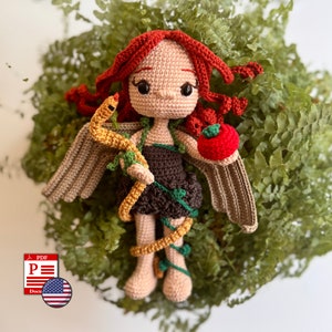 May include: A handmade crochet doll with red hair, holding a golden snake and a red apple. The doll has tan skin, a brown dress, and tan wings. Green vines wrap around the doll's body. The doll is set against a backdrop of green foliage.
