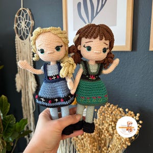 May include: Two crocheted dolls, one with blonde hair and a blue dress, the other with brown hair and a green dress. Both dolls have embroidered facial features and are holding hands.