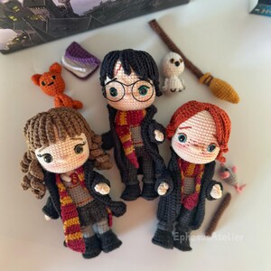 Pode incluir: Tr&ecirc;s personagens de Harry Potter feitos de croch&ecirc;: Harry Potter com &oacute;culos e uma echarpe vermelha e dourada, Hermione Granger com uma echarpe marrom e Ron Weasley com uma echarpe vermelha e dourada.