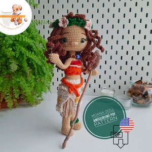 May include: A handmade crochet doll depicting a character with brown curly hair, wearing a floral crown, and a two-piece outfit in orange, white, and beige. The doll holds a wooden staff. The image includes text that reads "Moana Doll Amigurumi PDF Pattern."