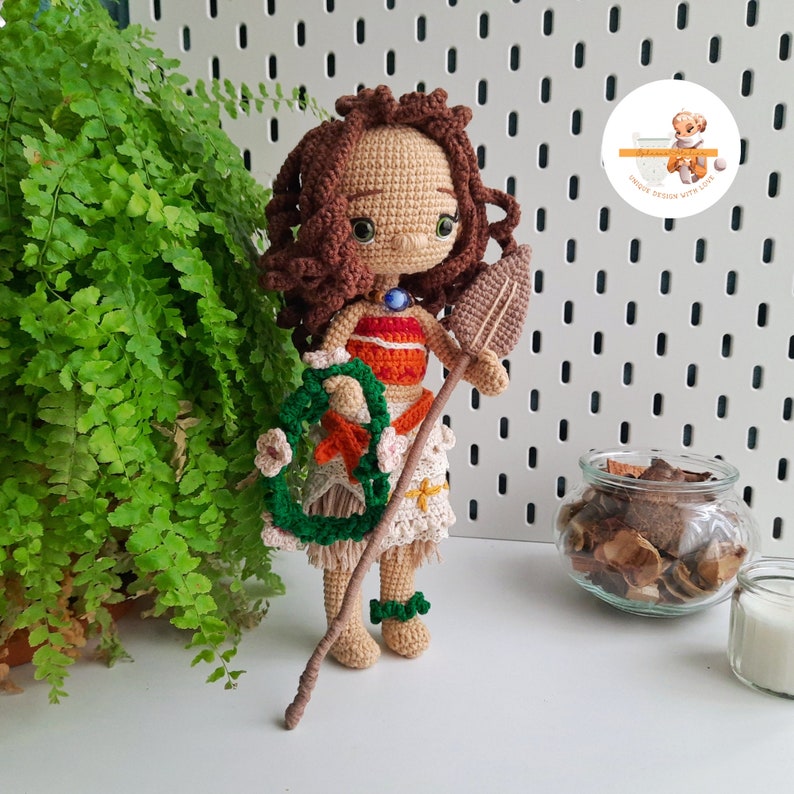 May include: A crocheted doll of a Polynesian princess, featuring brown curly hair, a tan complexion, and a detailed outfit. The doll holds a leaf-shaped paddle and a green wreath with flowers. The doll is approximately 10 inches tall.