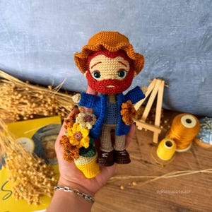 May include: Crocheted amigurumi doll of Vincent van Gogh, wearing a blue jacket and a brown hat, holding a pot of sunflowers.