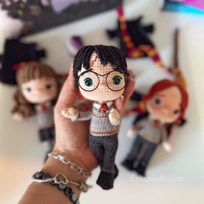 Crocheted Harry Potter doll with round glasses, a grey sweater, and a red and gold tie. The doll is holding a wand and has a lightning bolt scar on his forehead.