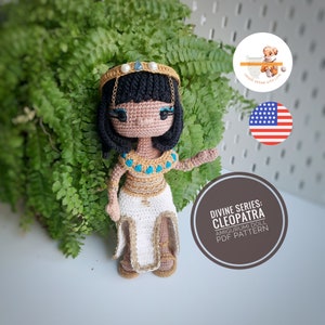 Pode incluir: Padrão de boneca amigurumi de crochê para Cleópatra, uma boneca amigurumi da série divina. A boneca está usando um vestido branco com detalhes dourados, um top azul e dourado e uma coroa dourada com uma gema azul. A boneca tem cabelo preto e olhos castanhos.