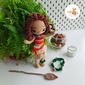 May include: A handmade crochet doll with brown hair, wearing a red and orange top, a white skirt, and green bracelets. Accessories include a green and white necklace and a brown staff. The doll is standing in front of a green plant.
