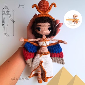 Könnte beinhalten: Häkelpuppe Amigurumi der ägyptischen Göttin Bastet. Die Puppe trägt ein weißes Kleid mit einem goldenen Gürtel und hat braunes Haar und blaue Augen. Die Puppe hält einen goldenen Stab und hat Flügel mit roten, blauen und weißen Federn. Die Puppe trägt eine goldene Krone mit einer Sonnenscheibe oben drauf.