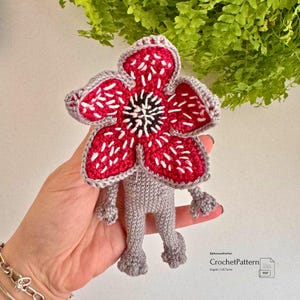 May include: A handmade crochet Demogorgon doll. The doll features a red flower-like head with white accents and a gray body with small legs and arms. The doll is held in a hand.