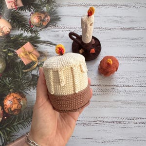 Könnte beinhalten: Gehäkelte Kerzen in Creme- und Brauntönen. Die Hauptkerze hat einen Wachstropfeneffekt und eine kleine Flamme. Eine kleinere Kerze und ein Kerzenhalter sind ebenfalls zu sehen.