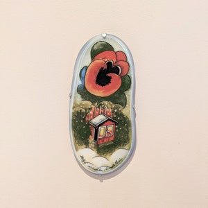May include: A ceramic tile with a painted image of a red house with a white roof and snow on the ground. A large red flower with a black center is in the upper part of the image. The tile is oval shaped and has a light blue background.