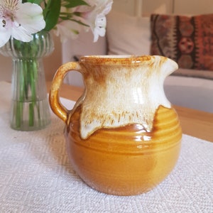 Puede incluir: Una jarra de cerámica marrón con un esmalte blanco. La jarra tiene un cuerpo redondeado y un asa.