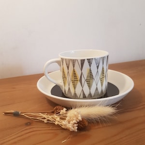 May include: A white ceramic coffee cup with a black and yellow geometric pattern. The cup is sitting on a white saucer with a black rim.