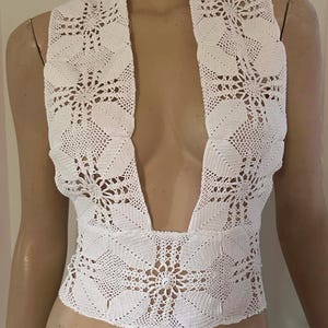 Könnte beinhalten: Weißes Häkeltop mit offenem Vorderteil und tiefem V-Ausschnitt. Das Oberteil hat ein Blumenmuster im gesamten Design. Die Häkelarbeit ist aufwendig, mit einem strukturierten Aussehen. Das Kleidungsstück wird auf einer Schaufensterpuppe präsentiert.