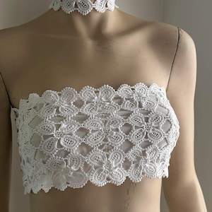 Puede incluir: Un top de tubo blanco sin tirantes y un collar gargantilla a juego. Ambas piezas están hechas de un delicado patrón de encaje floral. El top está diseñado para ser usado sin tirantes, y la gargantilla se coloca alrededor del cuello.